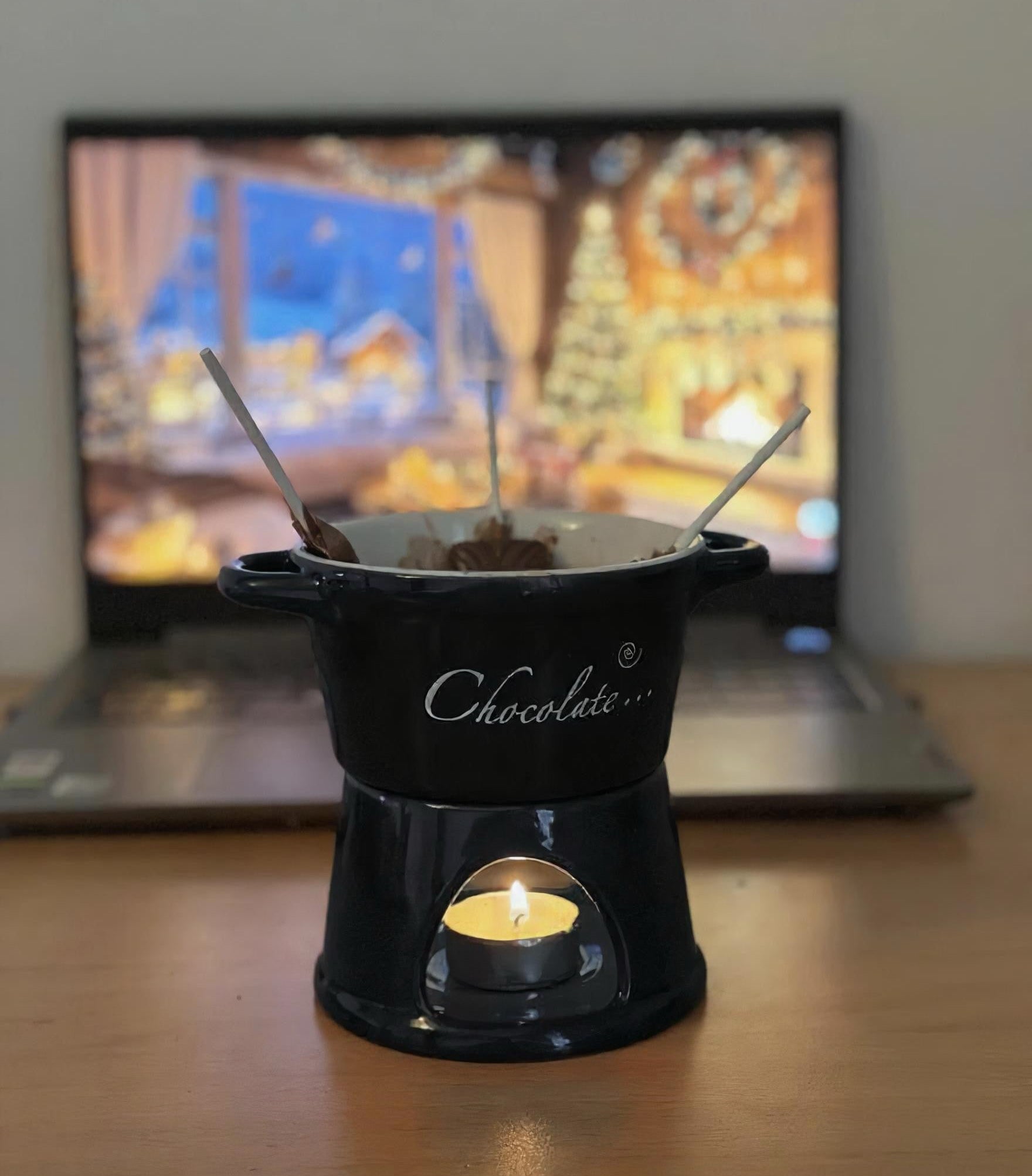 Black chocolate fondue pot on a wooden table with a blurred laptop in the background